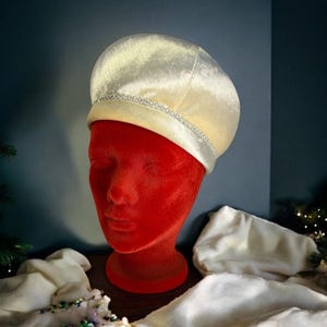 May include: A cream-colored satin beret featuring a delicate band of small, clear rhinestones encircling the crown. The hat has a smooth, lustrous finish and a rounded, structured shape that sits atop a red velvet display head form.