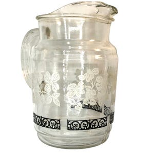 May include: Clear glass pitcher with a handle and a lid. The pitcher features a floral design in white and a black decorative band around the base. A classic piece for serving beverages.