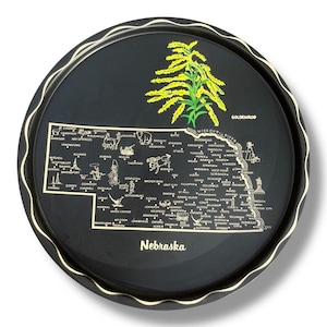 May include: A round black metal tray featuring a gold-colored map of Nebraska with various city names, small illustrations of local landmarks, and wildlife. The text Nebraska is printed at the bottom, and a yellow goldenrod flower is at the top.