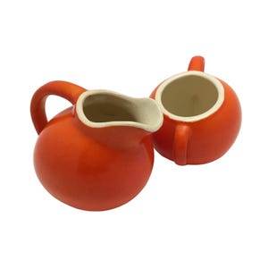 May include: A bright orange ceramic creamer pitcher with a curved handle and a rounded spout, paired with a matching orange ceramic sugar bowl featuring a small handle. Both pieces have a smooth, glossy exterior finish and a cream-colored interior.