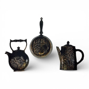 May include: Three decorative wall plaques in black with gold-toned highlights. The set includes a round pan with a fruit motif, a rounded teapot with a floral design, and a cylindrical teapot featuring an embossed botanical pattern on the surface.