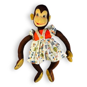 May include: A vintage stuffed monkey doll with a brown body, yellow ears and hands, and a smiling face. The monkey wears a white dress with a colorful print and an orange bodice. A charming collectible.