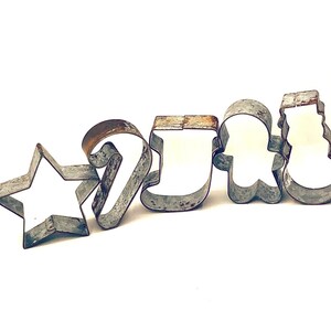 May include: A collection of vintage metal cookie cutters in various holiday shapes, including a star, candy cane, stocking, gingerbread man, and snowman. The cutters have a rustic, aged appearance with visible wear and tear, suggesting they are well-used.