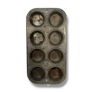 May include: A rectangular metal muffin pan with eight circular cups arranged in two columns. The surface shows signs of wear and oxidation. Embossed text on the center of the pan reads "CHICAGO" and "MADE IN USA" with additional markings.