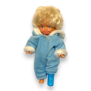May include: A vintage doll with blonde hair and blue eyes, wearing a light blue snowsuit with white trim. The doll has a zipper closure and a single blue boot. The doll's hands and one foot are visible.