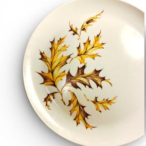 May include: A round, off-white ceramic plate featuring a decorative illustration of several oak leaves. The leaves are rendered in shades of golden yellow and deep brown, arranged in a cluster that extends across the surface of the plate.