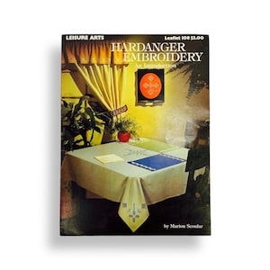 May include: A Leisure Arts instructional booklet titled Hardanger Embroidery: An Introduction, Leaflet 108. The cover features a yellow room with a table covered by a white cloth with embroidery, a green plant, and a wall hanging with a red design.