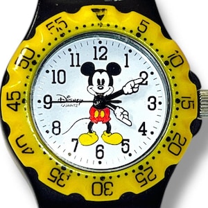 May include: A quartz wristwatch featuring a white dial with a graphic of Mickey Mouse, whose arms serve as the hour and minute hands. The watch has a bright yellow scalloped bezel with black minute markers and a black strap, with Disney printed.