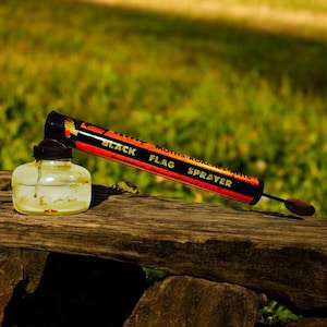 May include: A vintage Black Flag sprayer for moths and roaches, with a clear glass bottle and a black and red tube. The sprayer rests on a weathered wooden surface against a blurred green background.