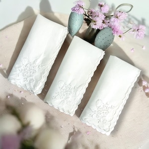 May include: Three white linen handkerchiefs with embroidered floral designs and scalloped edges are arranged on a light-colored surface. A vase of pink flowers and decorative eggs are in the background.