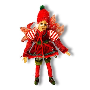May include: A whimsical Christmas elf figurine. The elf is dressed in a red velvet outfit with white striped sleeves, green accents, and gold bells. It has a red hat with a green pom-pom, and red glittery wings. The elf's face has a playful expression.