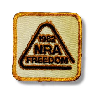 May include: A square embroidered patch with rounded corners, featuring a cream-colored background and a gold-colored border. Inside, a black triangular outline surrounds the black text 1982, NRA, and FREEDOM, all stitched onto the fabric.