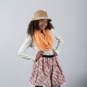 May include: A young person wearing a white long-sleeved shirt, a peach-colored scarf, a straw hat with a white band, a peach-colored skirt with a white eyeglass pattern, and pink shoes.
