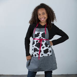 Op de afbeelding: Een jong persoon draagt een zwart shirt met lange mouwen, een schort met koeienprint met een rood lint, blauwe jeans en zwarte veterschoenen. Het schort heeft een grote zak aan de voorkant.