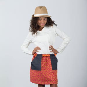 May include: A young person wearing a white long-sleeved shirt, a brown hat, a pink and gold patterned skirt with a denim pocket detail, and orange belt. The person is wearing pink canvas shoes.