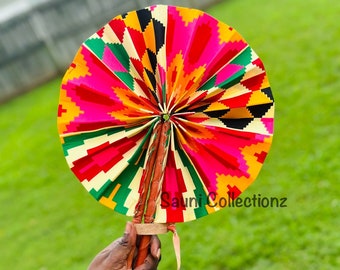May include: A colorful hand fan with a vibrant geometric pattern in shades of pink, red, yellow, green, and black. The fan is made of fabric and has a wooden handle. The text "Sauni Collectionz" is visible on the fan.
