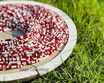 May include: A close-up of a red and white beaded necklace, coiled on a wooden plate. The necklace is made of small, round beads, and the colors are bright and vibrant. The plate is white and has a simple design. The background is a green grassy surface.
