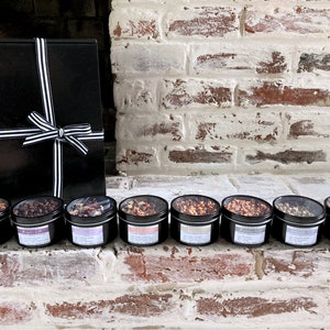 Puede incluir: Una caja de regalo negra atada con una cinta a rayas blancas y negras, junto a siete peque&ntilde;as latas negras llenas de varios t&eacute;s. Las latas est&aacute;n dispuestas en fila sobre una superficie de ladrillo.