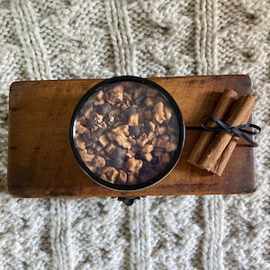 Puede incluir: Una caja de madera contiene un recipiente redondo lleno de frutas secas y especias, junto a un manojo de palitos de canela atados con una cinta negra. Los art&iacute;culos est&aacute;n sobre una superficie texturizada de color crema.