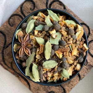 Puede incluir: Un cuenco negro lleno de mezcla de especias chai, que incluye an&iacute;s estrellado, vainas de cardamomo, clavos y c&aacute;scara de naranja seca. El cuenco est&aacute; sobre una tela marr&oacute;n estampada. Las especias son marrones, verdes y naranjas.