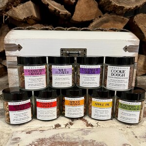May include: A collection of glass jars with black lids, each displaying a unique scent, including "Cranberry Orange," "Wild Flower," and "Cookie Dough." The jars are presented in front of a white wooden chest and a backdrop of stacked firewood.
