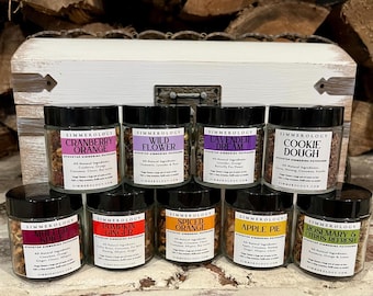 Puede incluir: Un surtido de frascos de vidrio con tapas negras, cada uno etiquetado con un aroma diferente, como "Cranberry Orange", "Wild Flower" y "Cookie Dough". Los frascos están dispuestos frente a un cofre de madera blanco y un fondo de leña apilada.
