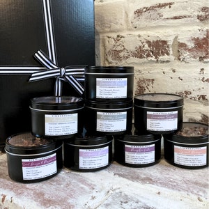 Puede incluir: Una colecci&oacute;n de botes de metal negro con popurr&iacute; para hervir a fuego lento, etiquetados con varios aromas como "Chamomile Fields" y "Sweet Orange & Peppermint". Una caja de regalo negra con una cinta a rayas se encuentra detr&aacute;s de los botes. Los productos se exhiben sobre una superficie de ladrillo.