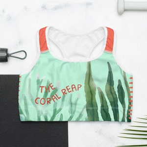 May include: A white sports bra with a green and blue watercolor design of seaweed. The bra has red straps and the text "THE CORAL REAP" is printed on the front.