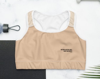 May include: A beige sports bra with a white mesh panel and a black text overlay that reads "NOTHING HAPPENS UNTIL SOMETHING MOVES".