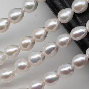 May include: A strand of white freshwater pearls, arranged in rows on a black surface. The pearls are smooth and have a pearlescent sheen.