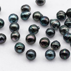 Könnte beinhalten: Eine Sammlung von dunkelgrünen und schwarzen, irisierenden Perlen, die auf einer weißen Oberfläche verstreut sind. Die Perlen sind in verschiedenen Größen und Formen, einige sind rund und andere leicht oval.
