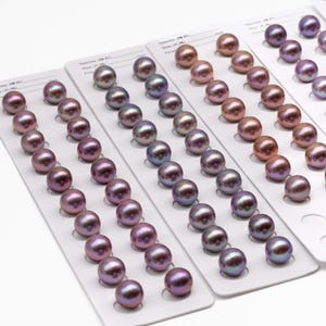 May include: A selection of iridescent pearls in pink, purple, and bronze hues, arranged on white cards. The round pearls have a smooth, reflective surface, displaying a spectrum of colours and subtle variations in tone.