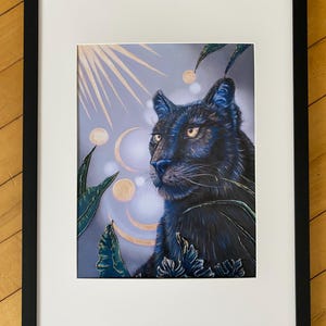 May include: A framed print of a black panther with a blue and gold celestial background. The panther is looking to the right and has a serious expression. The background is filled with stars, moons, and other celestial bodies.