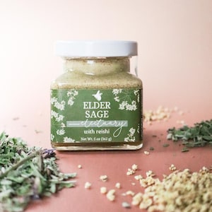 May include: A small glass jar with a white lid, filled with a light green powder. The jar has a green label with the words "Elder Sage Electuary with reishi." The label also includes floral illustrations and the net weight, 5 oz (142 g).