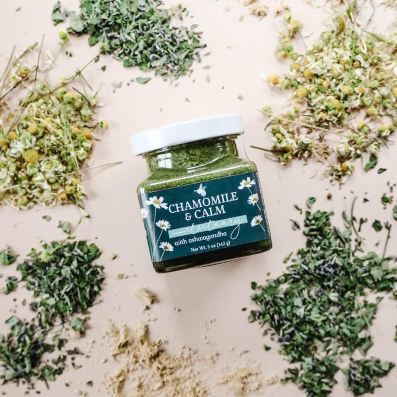 A square glass jar of Chamomile & Calm Herbal Honey Electuary with Ashwagandha. The label on the jar is a deep blue with a beautiful botanical design and the honey in the jar is a vibrant green color from powdered medicinal herbs. The jar sits against a light pink backdrop and is surrounded by small piles of dried herbs including chamomile tea, skullcap, anise hyssop, and dried powdered ginger root.