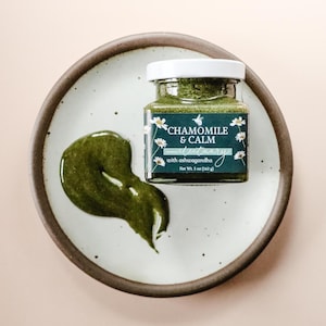 A square glass jar of Chamomile & Calm Herbal Honey Electuary with Ashwagandha. The label on the jar is a deep blue with a beautiful botanical design and the honey in the jar is a vibrant green color from powdered medicinal herbs. The jar sits on a white speckled plate with the brown clay rim and against a light pink backdrop.