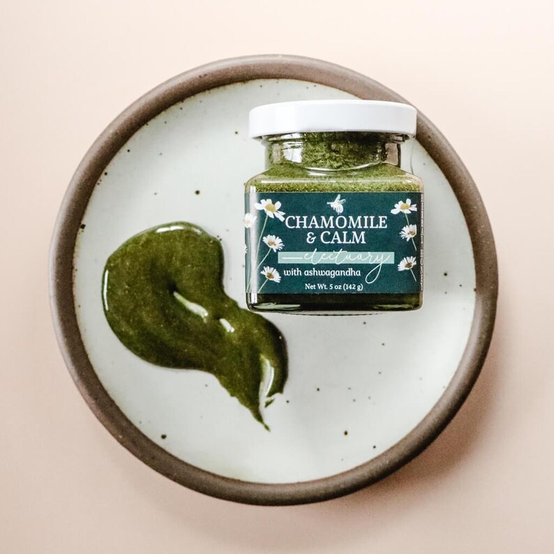 A square glass jar of Chamomile & Calm Herbal Honey Electuary with Ashwagandha. The label on the jar is a deep blue with a beautiful botanical design and the honey in the jar is a vibrant green color from powdered medicinal herbs. The jar sits on a white speckled plate with the brown clay rim and against a light pink backdrop.