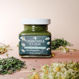 A square glass jar of Chamomile & Calm Herbal Honey Electuary with Ashwagandha. The label on the jar is a deep blue with a beautiful botanical design and the honey in the jar is a vibrant green color from powdered medicinal herbs. The jar sits against a light pink backdrop and is surrounded by small piles of dried herbs including chamomile tea, skullcap, and anise hyssop.