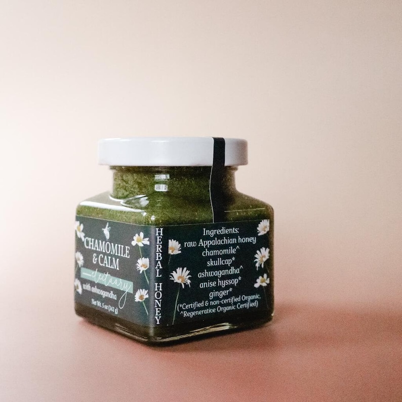 A square glass jar of Chamomile & Calm Herbal Honey Electuary with Ashwagandha. The label on the jar is a deep blue with a beautiful botanical design and the honey in the jar is a vibrant green color from powdered medicinal herbs. The labels contains the ingredients: raw Appalachian honey, chamomile, skullcap, anise hyssop, and ginger. All organic herbs. The jar sits against a light pink backdrop.