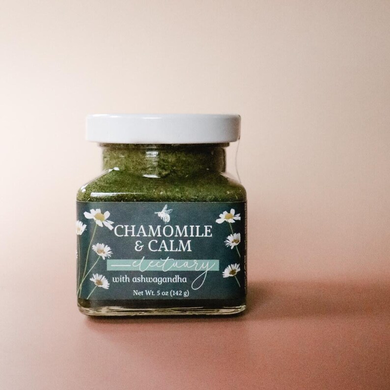 A square glass jar of Chamomile & Calm Herbal Honey Electuary with Ashwagandha. The label on the jar is a deep blue with a beautiful botanical design and the honey in the jar is a vibrant green color from powdered medicinal herbs. The jar sits against a light pink backdrop.