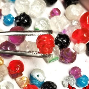 May include: A collection of small, translucent and opaque skull-shaped beads in various colors including red, black, blue, purple, and white. A pair of metal tweezers holds a single red skull bead. The text NIGHTMARECRAFTSTUDIO is visible.