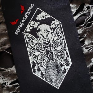 May include: Black rectangular patch with a white illustration of a moth with a skull-like head, set within a diamond shape. The patch includes the text "NIGHTMARECRAFTSTUDIO" and two red bat silhouettes. The design features a forest and a full moon.