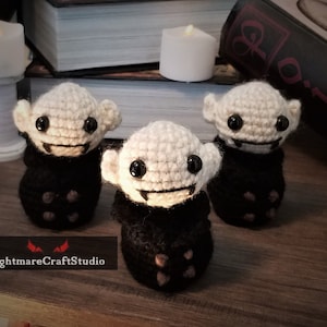Könnte beinhalten: Drei handgefertigte Häkelpuppen in Vampirform mit schwarzen Knopfaugen, cremefarbener Haut und schwarzen Umhängen. Die Puppen haben kleine Fangzähne und spitze Ohren. Die Puppen stehen auf einer Holzoberfläche mit Kerzen und Büchern im Hintergrund. Der Text "NightmareCraftStudio" steht unten.