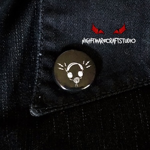 May include: A black button with a white graphic of headphones with sound waves coming out of them. The button has a silver metal backing.