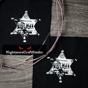 Könnte beinhalten: Zwei schwarze Stoff-Patches mit weißen gestickten Sternformen. Die Sternformen haben das Wort "HOOLIGAN" darin geschrieben. Die Patches befinden sich auf einer Holzoberfläche.
