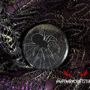 May include: A black and white button with a spider silhouette on a web. The button is on a purple and black lace background.