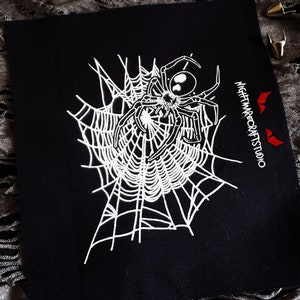 Peut inclure: Patch en tissu noir avec une araignée brodée en blanc et le texte "NIGHTMARECRAFTSSTUDIO" en blanc.