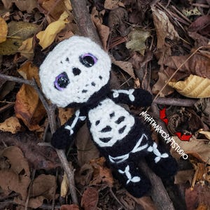 May include: A handmade crochet skeleton doll with a white head and black body, detailed with white bone accents. The doll has purple eyes and is lying on a bed of fallen leaves and twigs. The doll is a Halloween or Day of the Dead themed item.
