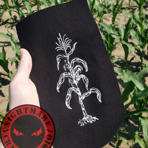 May include: A rectangular black fabric patch featuring a white line art illustration of a corn stalk with roots. The patch includes a circular logo in the bottom left corner with the text NIGHTMARE CRAFT STUDIO and two red eyes.