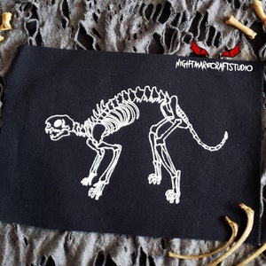 May include: White embroidered skeleton of a cat on a black fabric patch. The patch has the text "NIGHTMARECRAFTSTUDIO" in white letters.